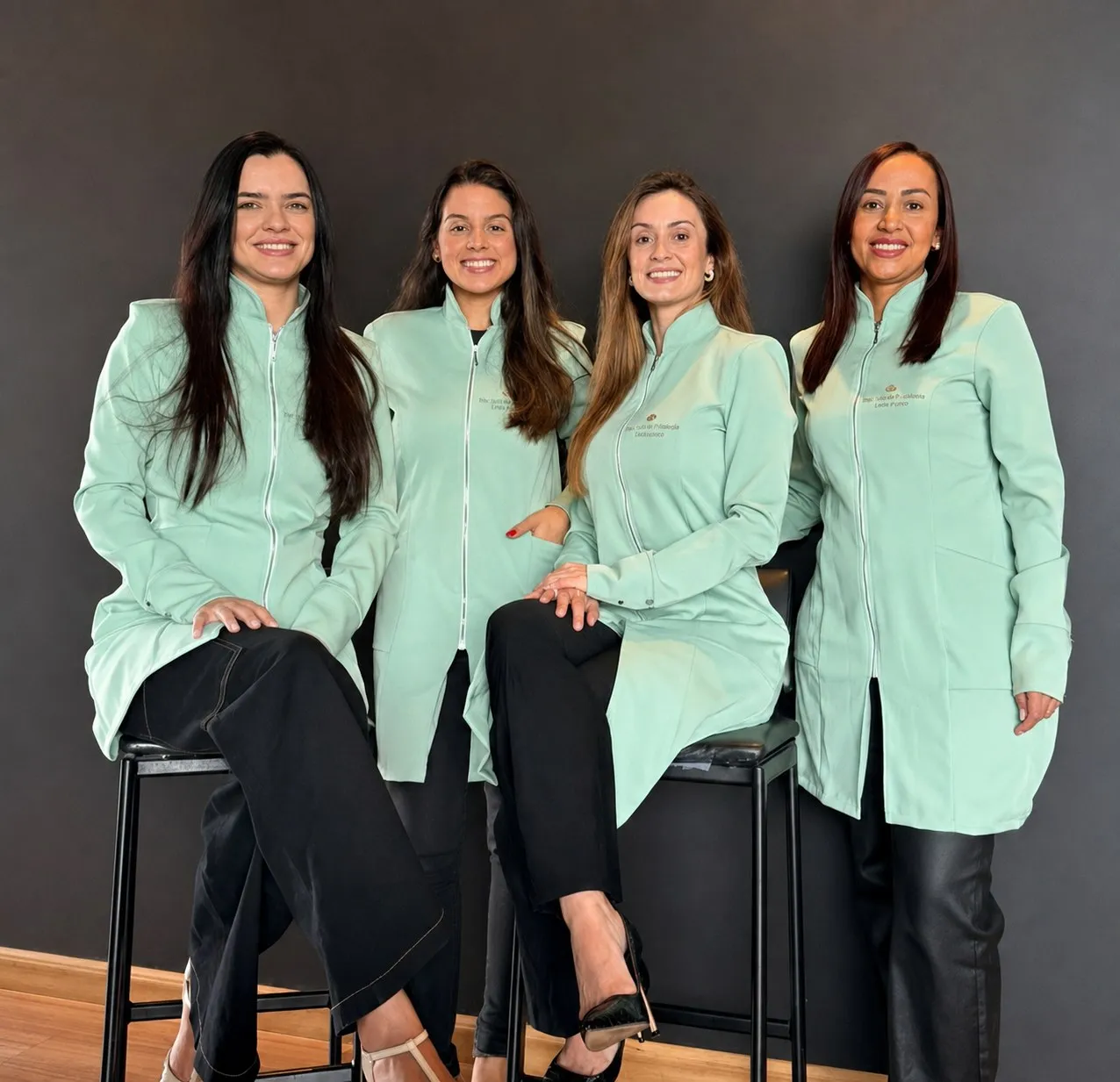 As quatro cirurgiãs-dentistas da equipe de home care, de jaleco verde-menta, em frente a uma parede cinza-chumbo